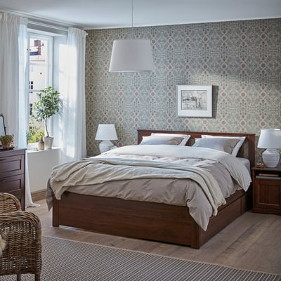 Dark wood bed, light linens, patterned wallpaper, white lamps, plant, wicker chair, large window.