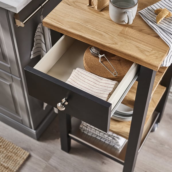 Wooden kitchen cart with open drawer showing utensils, blue mug, and striped cloth.