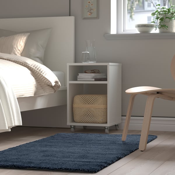 Modern bedroom with bed, white STOENSE rug, wooden stool, and side table with books and a basket.