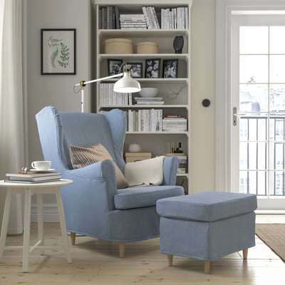 Cosy corner with blue/white striped STRANDMON chair & ottoman. Bookshelf, lamp, window add to serene atmosphere.