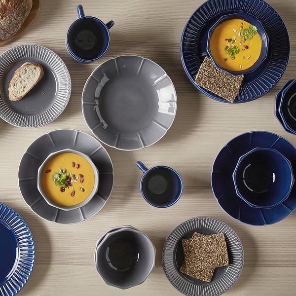 Several STRIMMIG plates and cups on a table, dark blue and grey colours, modern crafted feel, some with soup, bread, and garnish.