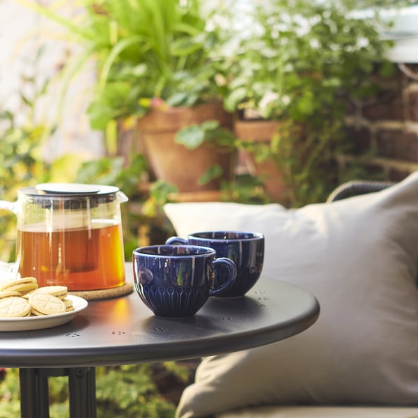 Small outdoor table with tea pitcher, two blue mugs, and cookies. Potted plants in the background.