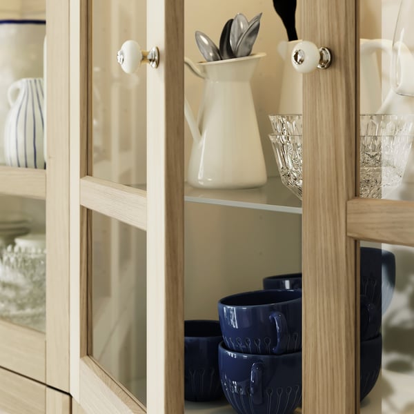 Wooden cabinet with glass doors holding white pitchers, clear glasses, and blue ceramic bowls. Mugs are uniquely patterned and medium-sized.