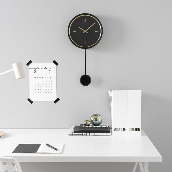 Minimalist desk setup with black pendulum clock, january wall calendar, books, office supplies, and white chair.