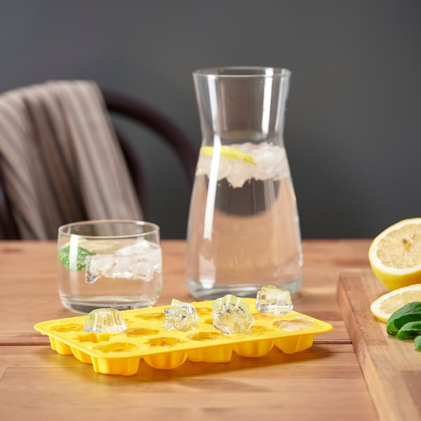 Bright yellow ice cube tray with diamond-shaped ice cubes. Glass with ice, water jug, and sliced lemons on wooden table.