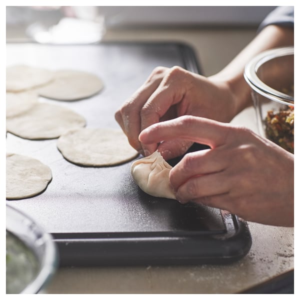 Hands fold dough on a black elliptical chopping board with round dough pieces nearby.