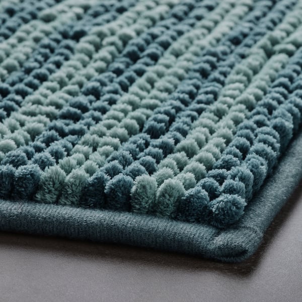 Displays a soft, teal bath mat with a textured, loopy surface. The fabric appears plush and inviting, showcasing its high-quality material.