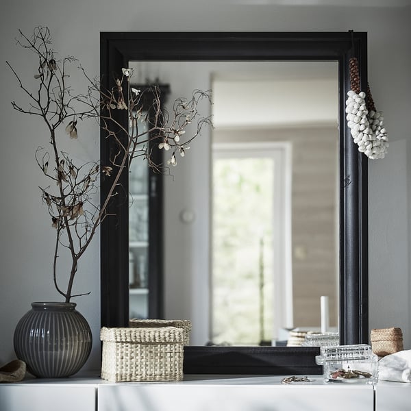 Black rectangular wall mirror, framed, styled with decorative branches, white woven basket, and grey vase on a white shelf.
