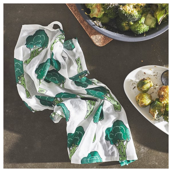 White towel with green broccoli prints lies on dark surface, surrounded by plates of roasted broccoli.