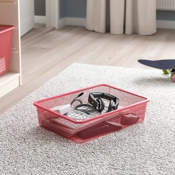 Red mesh storage box on carpeted floor, containing headphones and cables. Part of TROFAST storage system for organising accessories.