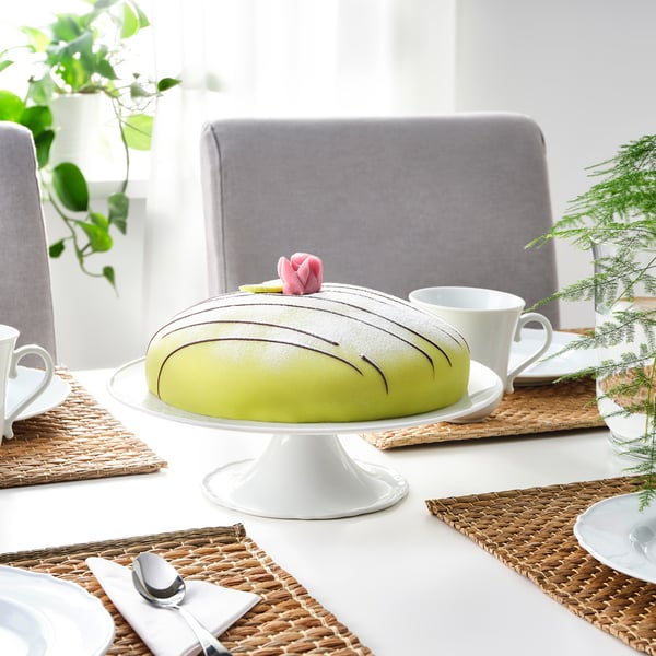Cake stand with green cake, white cups, on table with woven place mats, chairs.