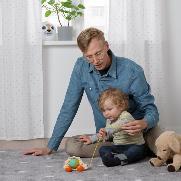 Person plays with toddler and UPPSTÅ hedgehog toy. Boy pulls string, developing motor skills. Stuffed animal nearby, white curtains in background.