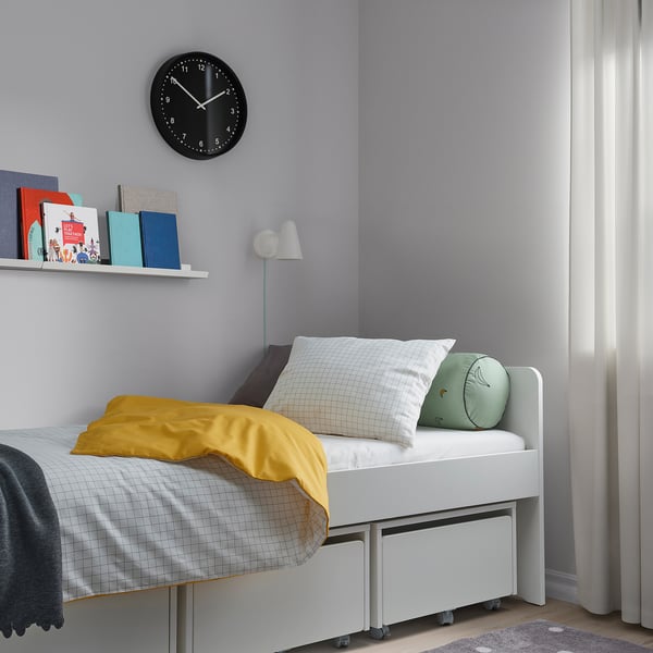 Small, colourful bed with VÄNKRETS yellow duvet, two pillows, and bookshelf with books and clock.