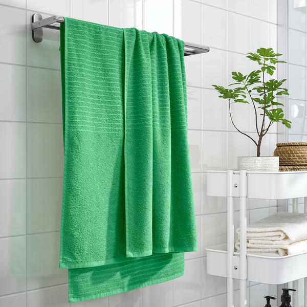 Green VÅGSJÖN towel on chrome bar, above white towels, vibrant against tiles, plant adds nature.