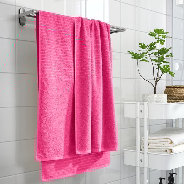 Bright pink VÅGSJÖN towel hangs on wall rack, surrounded by white tiles. Its thick, soft cotton makes it larger than adjacent folded towels on stand.