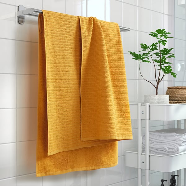 Bright yellow VÅGSJÖN towel on rod, soft & absorbent, 400 g/m², fits well in bathroom, hangs beside potted plant.