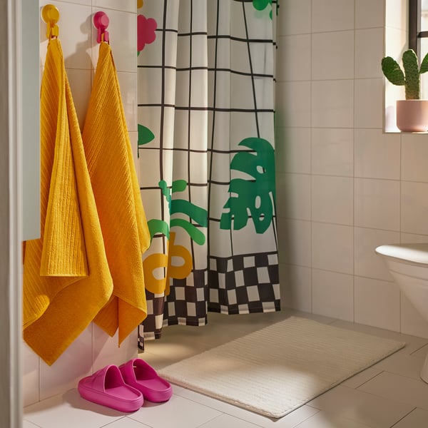 Bright bathroom with yellow VÅGSJÖN towels on colourful hangers, modern shower curtain, pink slippers, and potted plant.