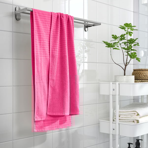 A bright pink VÅGSJÖN towel hangs on a silver rod against white tiles. Nearby, a shelving unit holds folded towels and a green plant.
