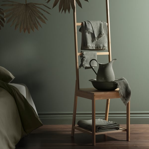 Modern bedroom with green walls, wooden chair holding folded light green vallasan towel, and decorative pitcher.