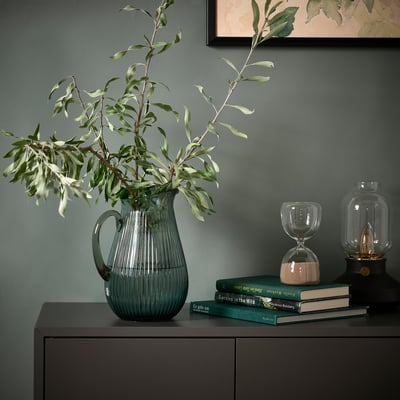 Green VARDAGEN glass jug with branches on dark cabinet, books, sand timer, green wall, painting.