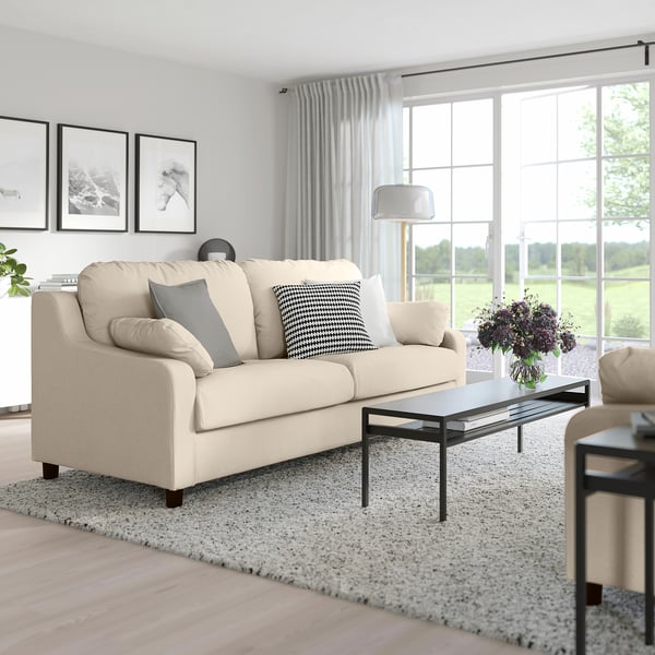 Cosy living room with beige VINLIDEN sofa, black-framed glass table, curtains, rug, and art. Sofa is large and inviting.