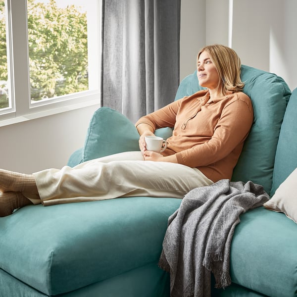 A person relaxes on a teal VINLIDEN sofa, holding a coffee cup, with a grey blanket beside the person. The person wears an orange sweater.