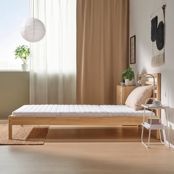 Minimalist bedroom with beige ÅNNFJÄLLET bed, side table, and decor. Large window, curtains, pendant light, plants, and wall hanging.