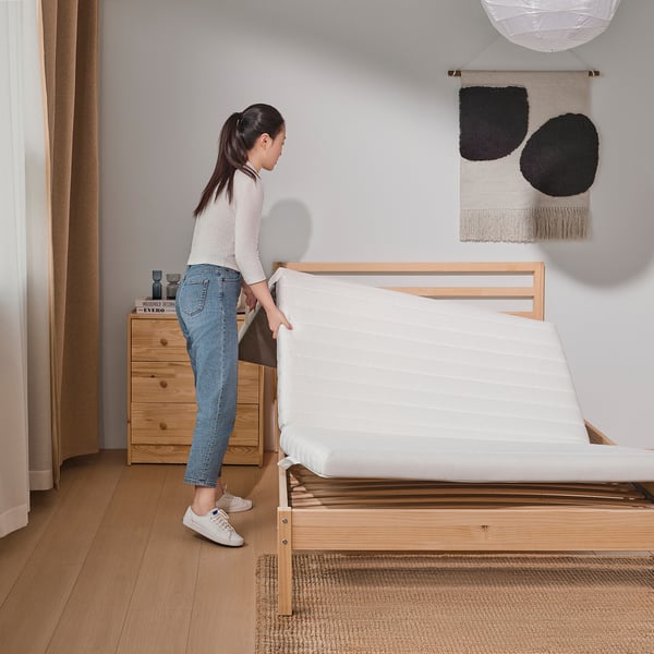 Person making bed with natural, foam-topped mattress on wooden frame in modern room.