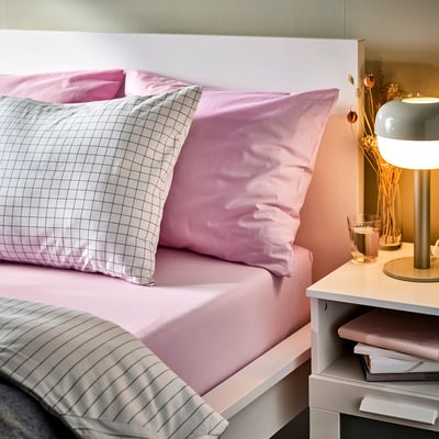 Pink bed with DVALA sheet, pink pillow, chequered pillow, striped blanket. Bedside table with lamp, glass, dried flowers.