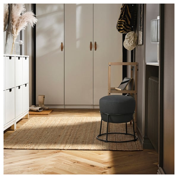 Modern entryway with dark grey EMMISLÖV pouffe, jute rug, stool, and coat rack.