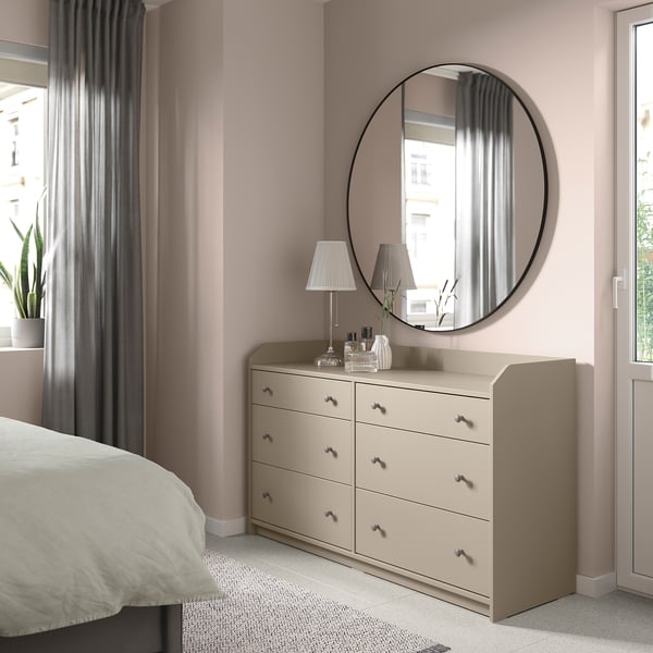 Bedroom with beige HAUGA chest, round mirror, lamps, plants, and curtains.