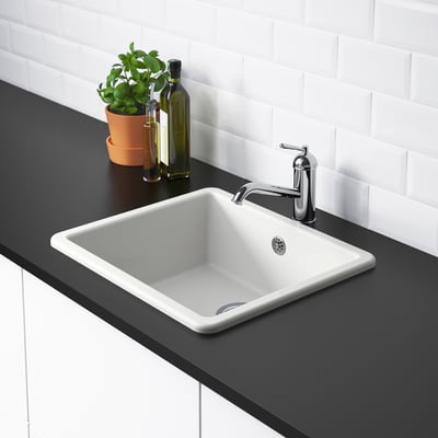 A white kitchen sink with a silver tap on a dark countertop. Two bottles and a plant are beside it.