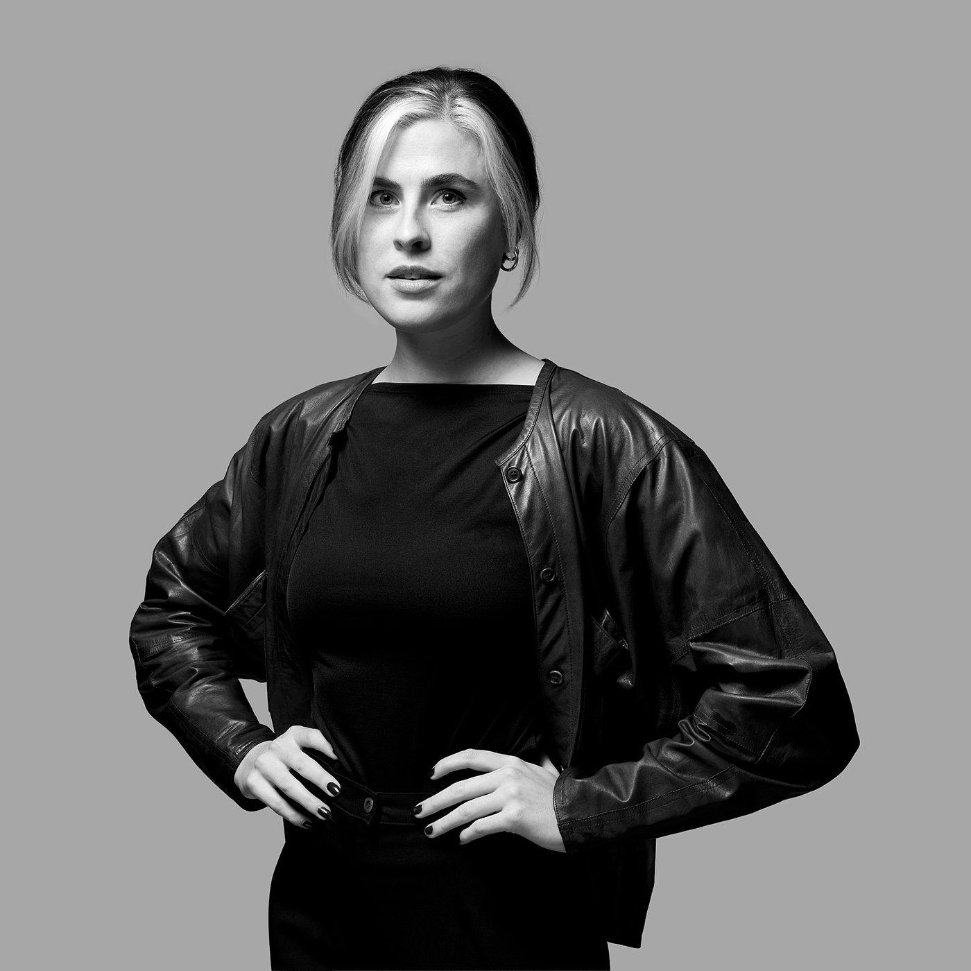 Black and white photo of the designer in a leather jacket and dark top, posing confidently with hands on hips.