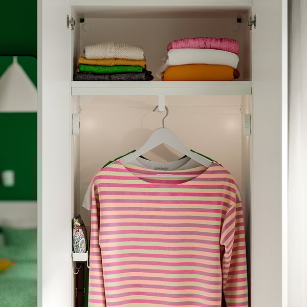 A closet with shelf and hanging rod. Pink and yellow striped shirt hangs on hanger. Folded clothes on top shelf.