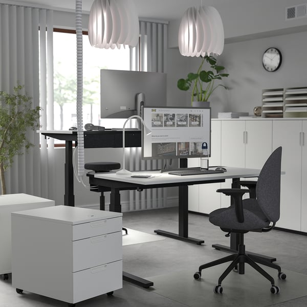 Modern minimalist office with mitzon adjustable desk featuring dual monitors, sleek black chair, and white drawers. Large windows and hanging lights.