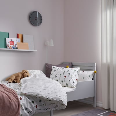 Kids bed with NATTSLÄNDA lavender duvet, floral patterns. Against lilac walls, with wall clock, bookshelf, lamp, and teddy bear.