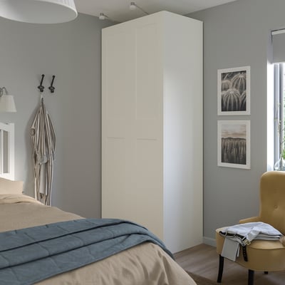 A minimalist bedroom with a large white PAX wardrobe. The room includes a bed with a dark blue blanket, a yellow chair, and frames.