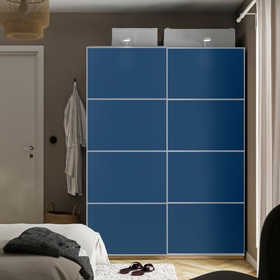 Modern bedroom with tall blue PAX wardrobe. Sliding doors, hanging clothes, and shoes on floor. Bed and door visible.