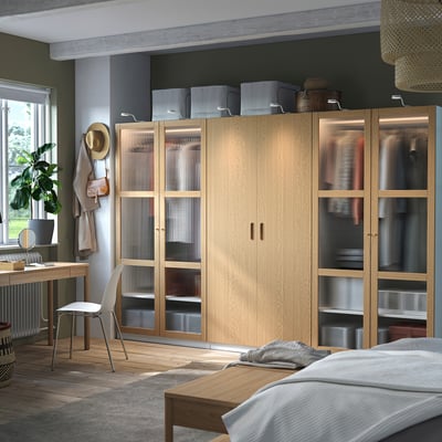 Modern bedroom with light wood PAX wardrobe, white bed, wooden desk, and plants.