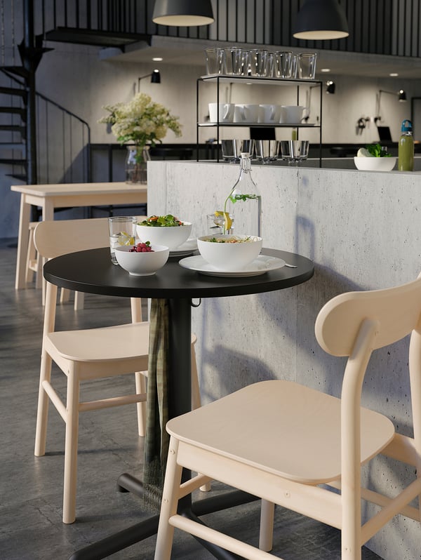 Modern dining area with white minimalist chairs and black round table. Table holds white bowls and a vase with greenery.