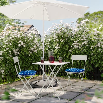 Outdoor setup with white foldable table and chairs, blue cushions, under a white umbrella, surrounded by blooming white flowers.