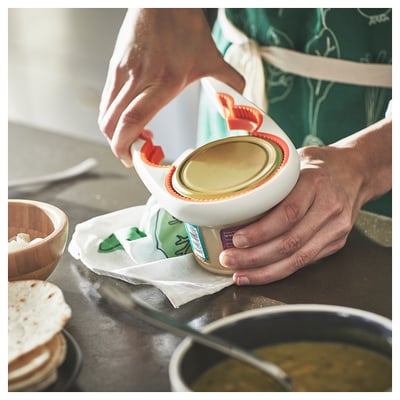 Person uses a white UPPFYLLD jar opener, squeezing it tightly to open a jar. A green apron and various kitchen items are visible in the scene.