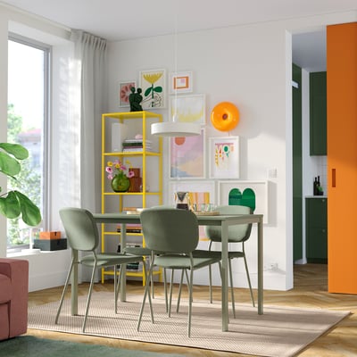 Bright dining space with modern yellow shelving, sleek grey chairs, and an orange door. Artwork and plants decorate the room.