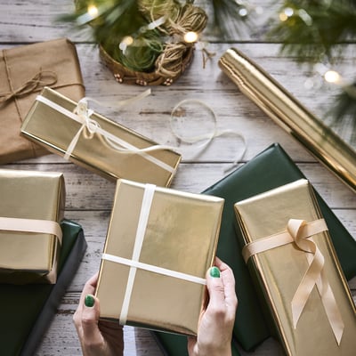 Hands wrapping gifts with golden paper and white ribbon on rustic table with christmas decorations.