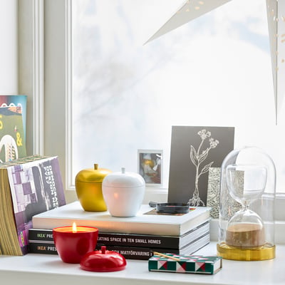 A shelf with VINTERFINT red apple candle, small, between books, lit. Bright red, tin steel, realistic shape.