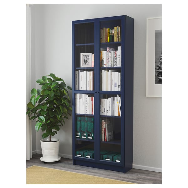 Blue BILLY bookshelf with glass doors displaying various books and items in a living area, with a potted plant beside it.