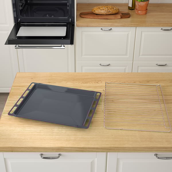 Black baking tray and wire rack rest on wooden countertop near open oven with white cabinets.