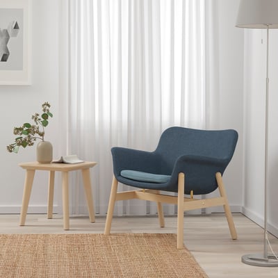 Blue VEDBO armchair with cushioned seat and curved back next to a light wood side table and beige floor lamp on a patterned rug.