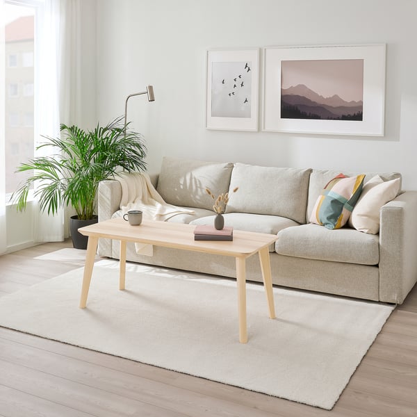 Bright, minimalist living room with KNARDRUP white rug, light gray sofa, wooden coffee table, and wall art. Plants add greenery.