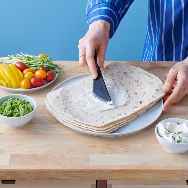 Person spreading cream on flatbread with knife. Colorful veggies and herbs nearby.